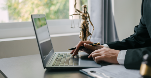 The hands of a man in a suit typing on a laptop at a desk. A statue of Lady Justice sites on the desk behind the laptop.