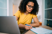 A woman is sitting at her desk behind an open laptop and reviews a checklist with pen and paper.