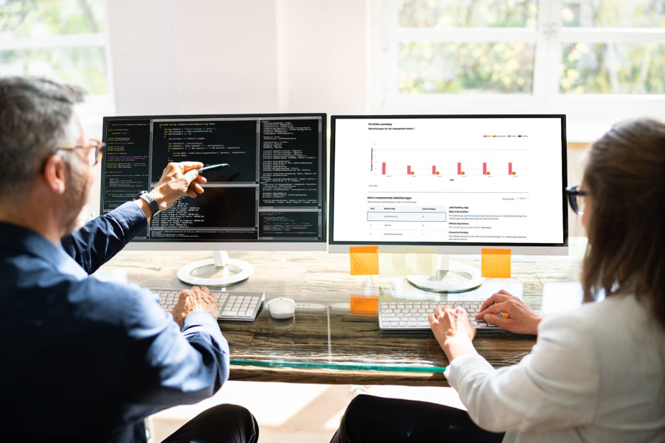 Two professionals collaborating in an office, with one explaining data on a screen and the other typing on a computer.