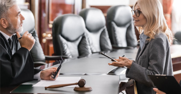 A female lawyer with blonde hair speaks with a gray-haired male judge in a legal court. A gavel sits on the table between them.