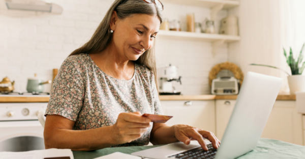 A smiling woman uses her credit card to make a purchase on a laptop. She has long gray hair, wears a floral shirt, and sits at a kitchen table.