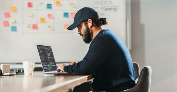 A male developer writes code on a laptop while seated in a modern office. He has a dark beard and wears a baseball cap and earbuds. Colorful sticky notes are posted on a whiteboard behind him.