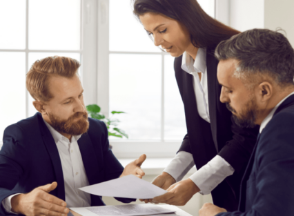 Three lawyers review a legal document around a table in an office space. One of the lawyers, a woman with dark hair, holds the document while two male lawyers look at it with frustrated expressions.