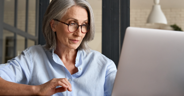 A woman uses a laptop computer. She has gray hair, wears glasses, and has a thoughtful expression.