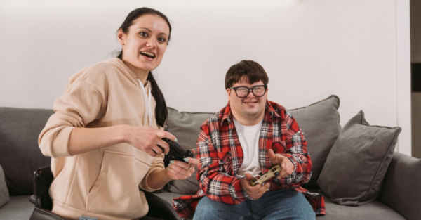 A young woman using a wheelchair and a young man with Down's syndrome joyfully play a video game together. The young man is seated on a gray couch.
