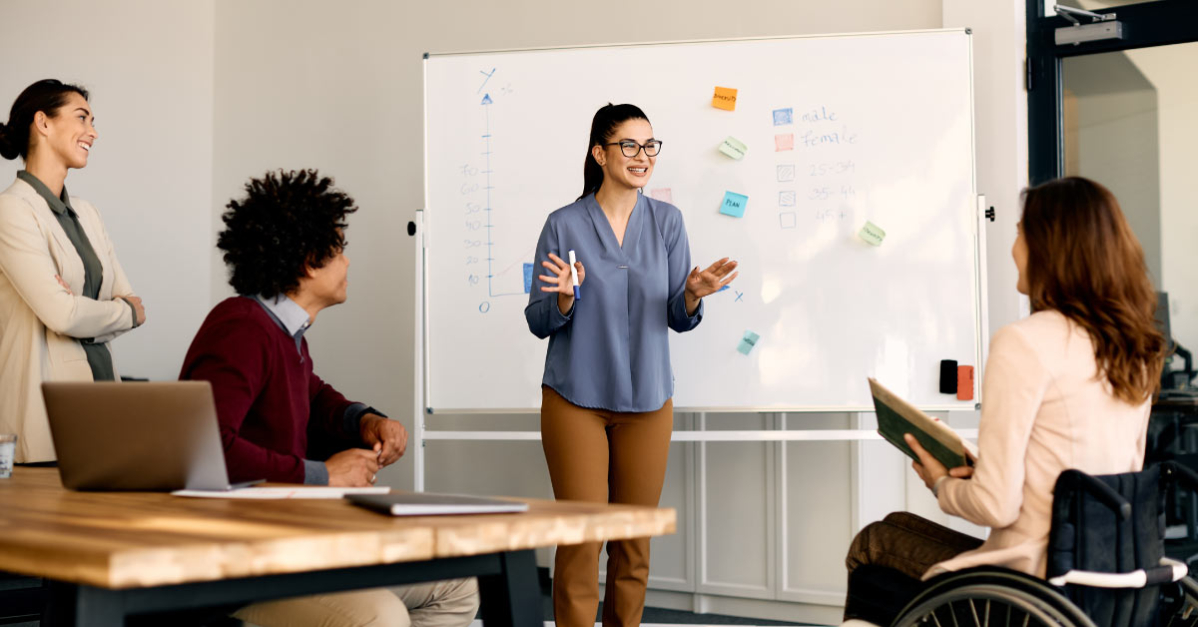 A young businesswoman standing in front of a white board speaks excitedly to three teammates, one of whom is using a wheelchair