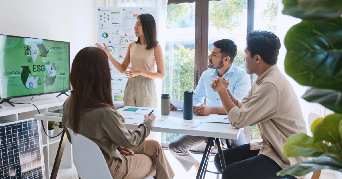 A professional woman with dark hair gives a presentation on ESG to three other professionals seated around a table in an office